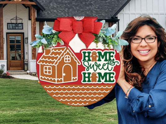 A smiling woman with long brown hair and glasses holds a round Home Sweet Home sign decorated with a gingerbread house, greenery, and a red bow, standing in front of a modern farmhouse-style home.