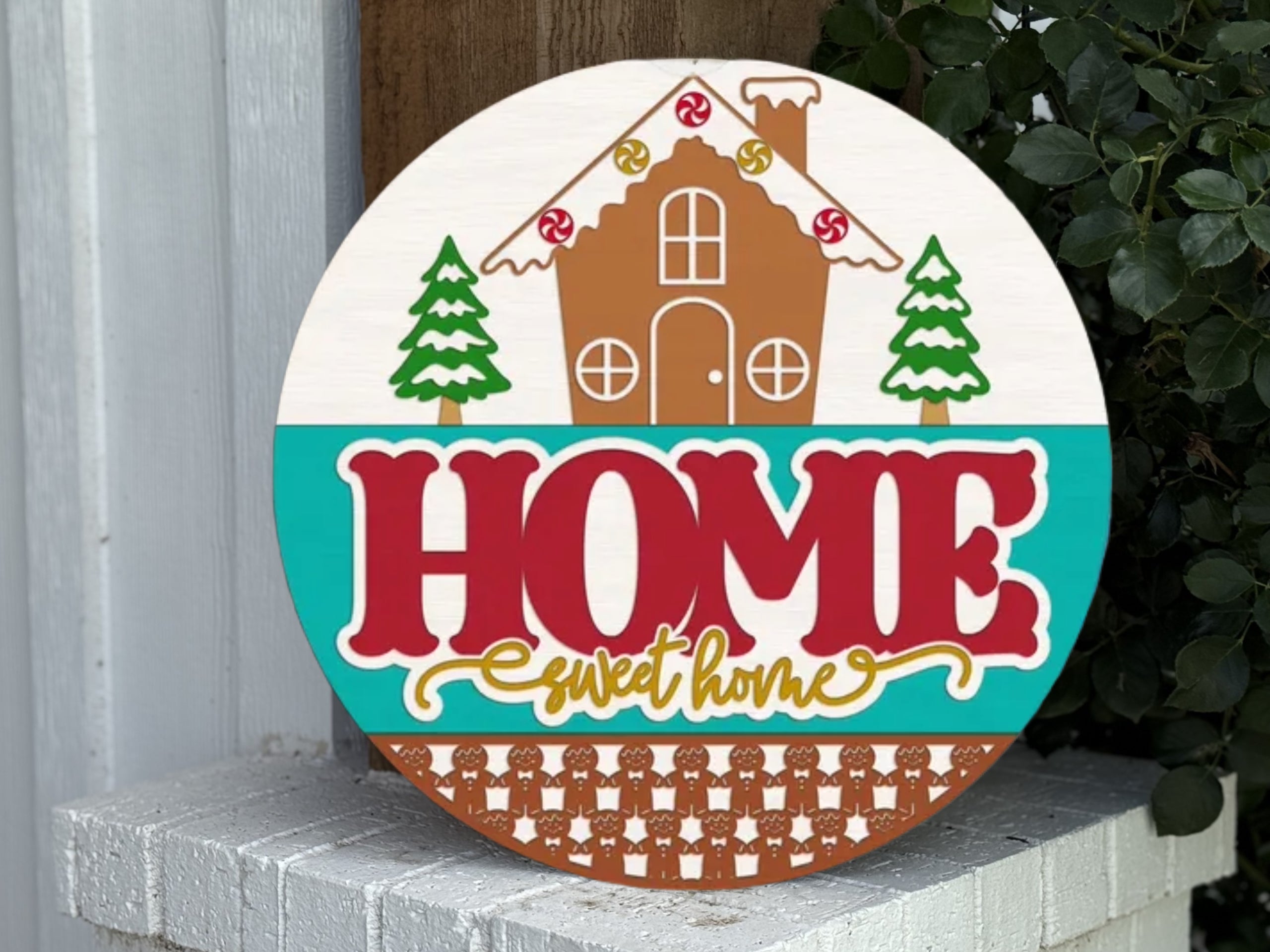 A decorative round sign with a gingerbread house, candy decorations, pine trees, and the words “HOME sweet home” in bold red and yellow letters, displayed on a white brick ledge with greenery nearby.