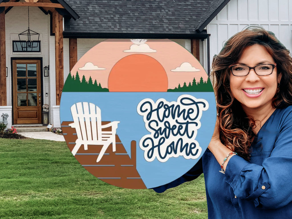 A woman smiles while holding a circular sign that reads Home Sweet Home with a sunset, lake, trees, and an Adirondack chair illustration, standing in front of a modern farmhouse.