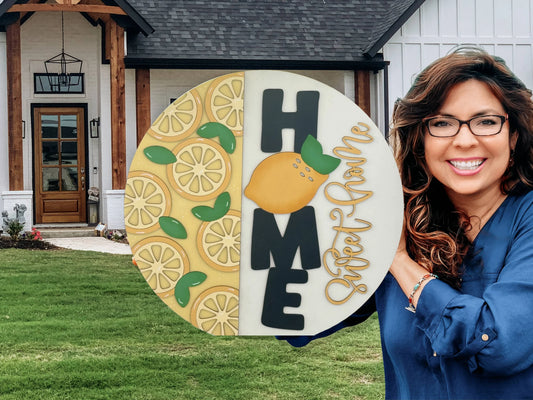 Woman with long brown hair and glasses, wearing a blue top, smiles while holding a large round sign. The sign reads HOME Sweet Home with lemon graphics on the left. A modern house with a wooden door and large windows is in the background, set in a well-kept lawn.