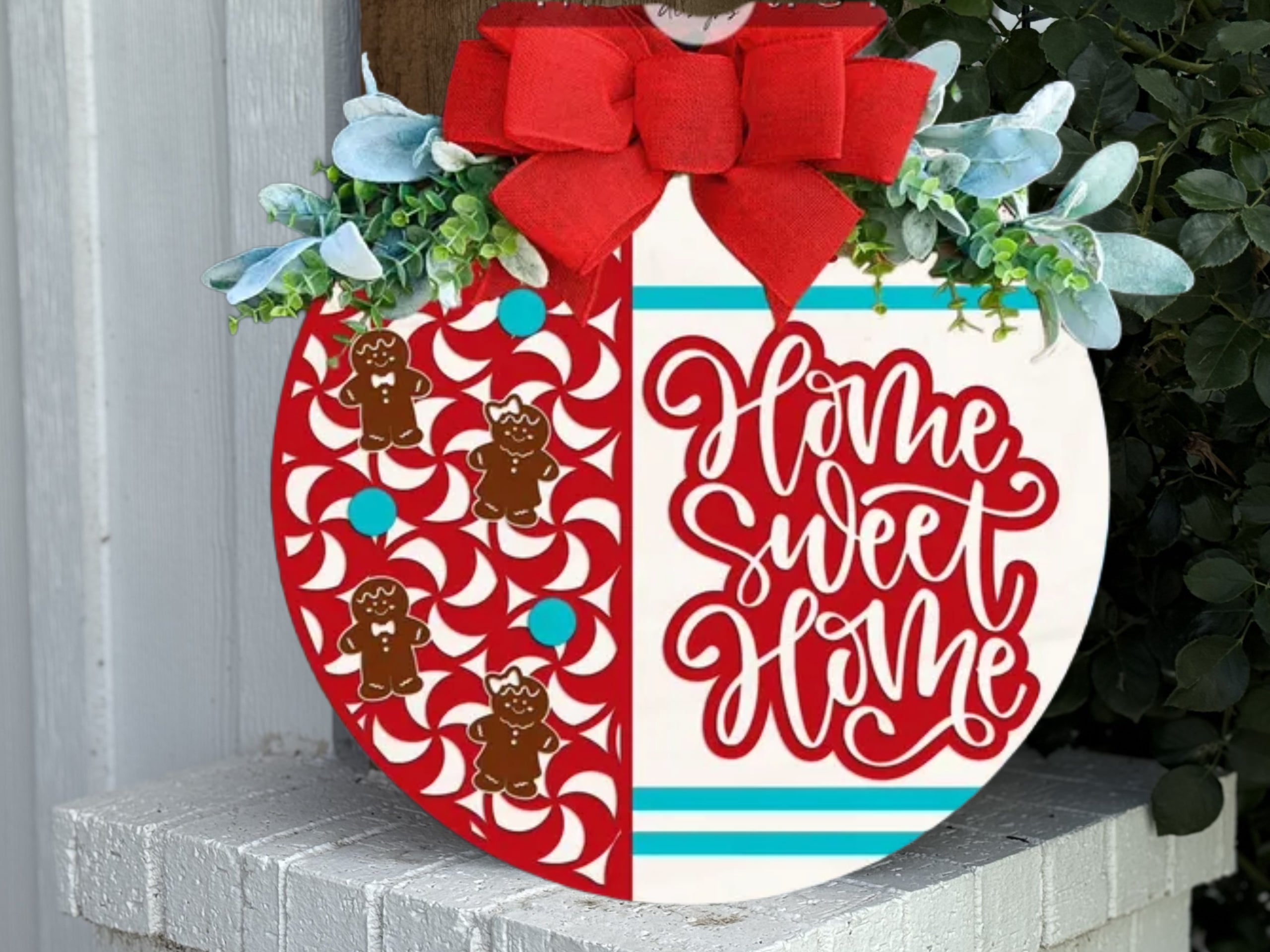 A festive round sign decorated with red and white peppermint patterns and gingerbread men on the left, Home Sweet Home in red script on the right, topped with a red bow and greenery, displayed on a white brick ledge.
