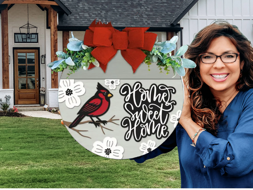 A smiling woman with long brown hair and glasses holds a decorative Home Sweet Home sign featuring a red cardinal, flowers, and a red bow, standing in front of a modern house with a green lawn.
