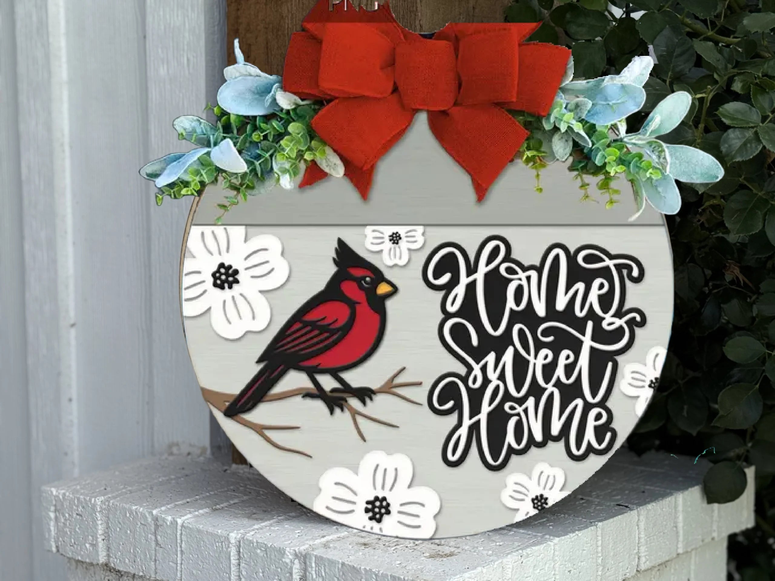 A round wooden sign with a red cardinal, white flowers, and the words Home Sweet Home in black script. It is decorated with a red bow, greenery, and is displayed on a white brick surface.