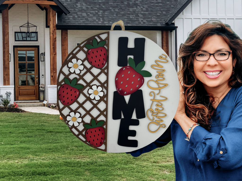 A smiling woman with long brown hair and glasses holds a round Home Sweet Home sign decorated with strawberries and flowers, standing in front of a modern house with wood and white exterior.