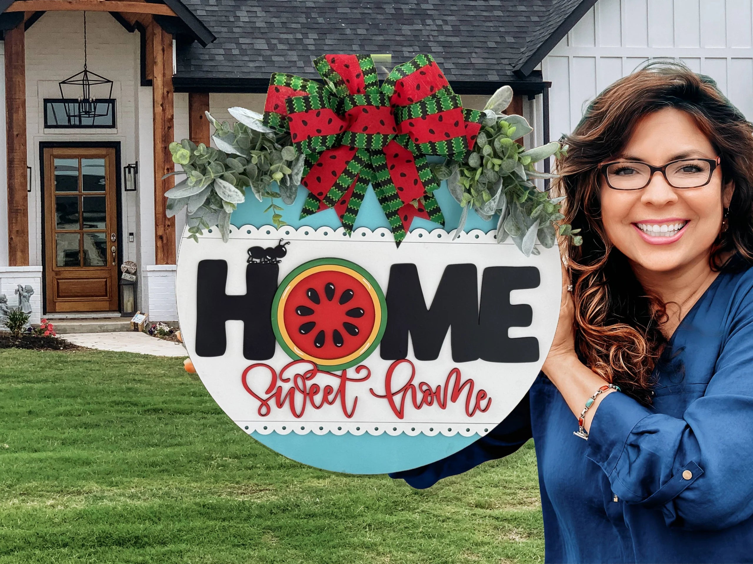 A smiling woman with wavy hair and glasses holds a circular sign that reads HOME Sweet Home. The sign features a watermelon slice and a red and green bow. She stands in front of a modern house with a wooden door and large windows, set against a manicured lawn.