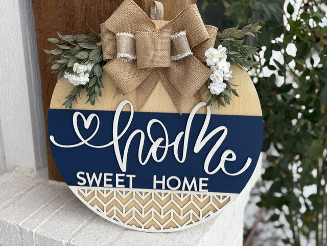 A round wooden sign hangs on a wall, featuring the words home sweet home in white cursive and block letters on a blue background. The top is adorned with a burlap bow and small white flowers surrounded by green leaves. The bottom features a zigzag pattern in beige and white.