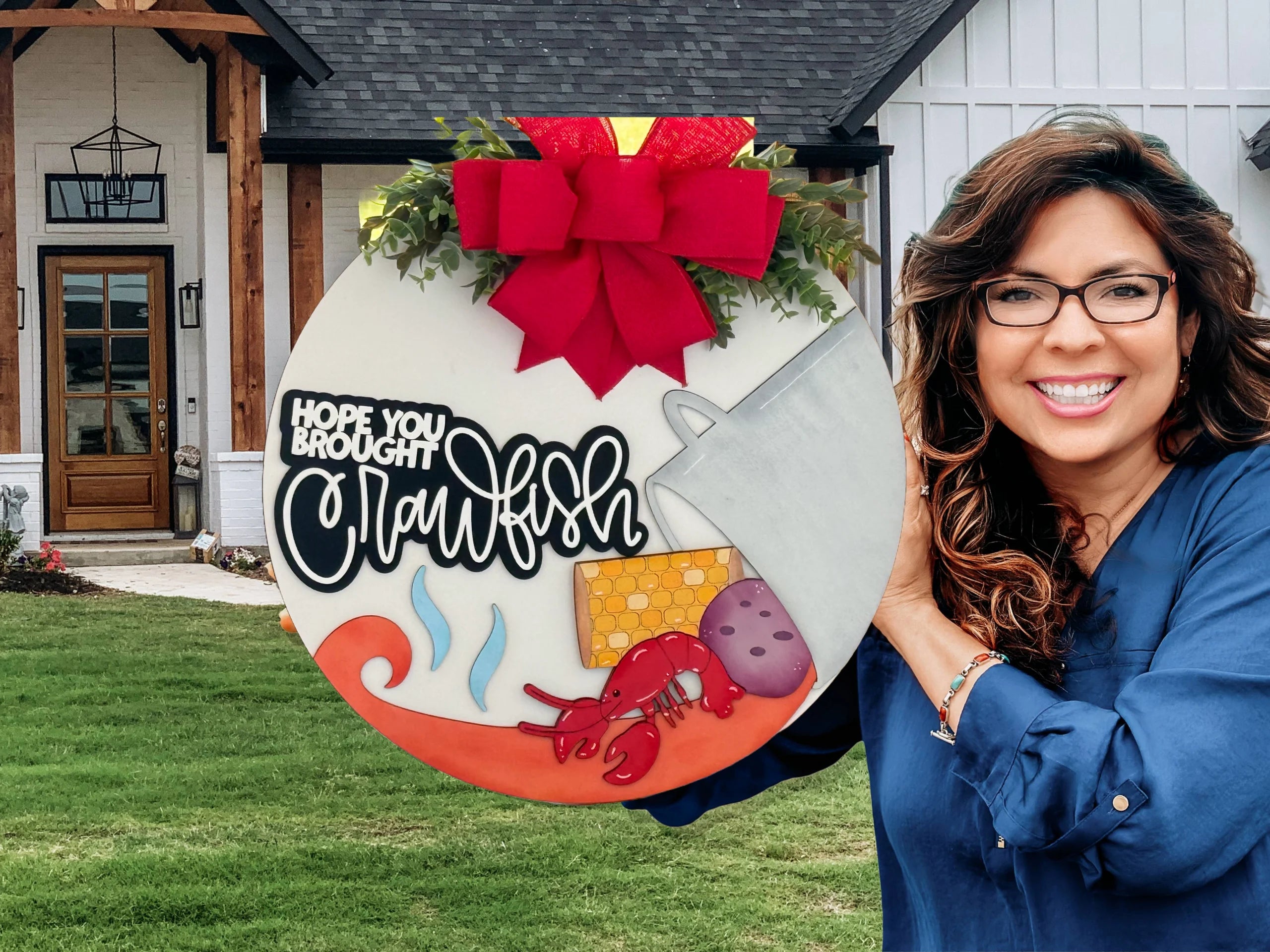 A smiling woman with wavy brown hair and glasses holds a round sign with a red bow that reads, “Hope you brought crawfish.” The sign features images of a crawfish, corn, potatoes, and a pot. A modern white house with a wooden porch is in the background.