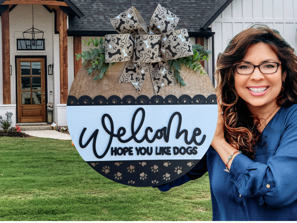A smiling woman holding a round sign that says Welcome, hope you like dogs with a paw print design and a decorative bow, standing in front of a modern house.
