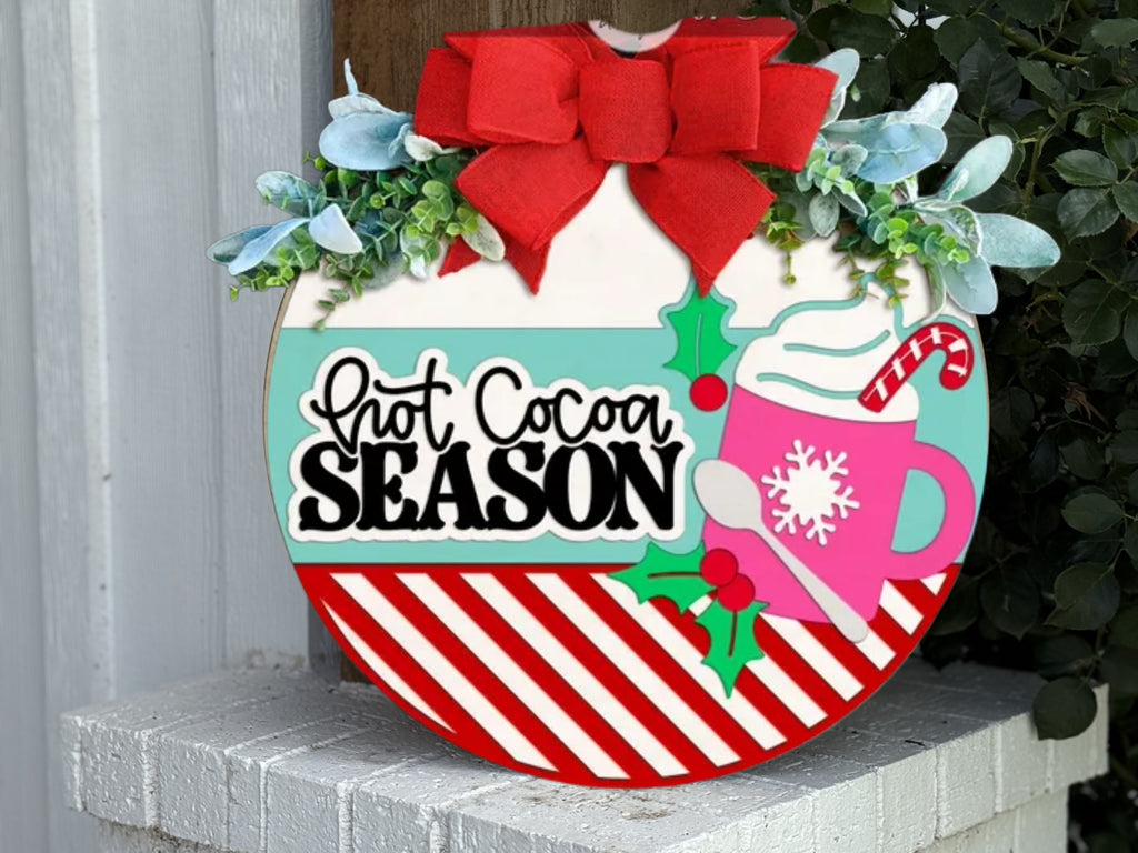 A round holiday sign with a red bow, green leaves, and the text “Hot Cocoa Season.” It features a pink mug with a snowflake, whipped cream, and a candy cane, plus red and white diagonal stripes at the bottom.