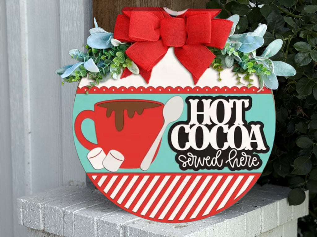 A decorative sign with a red cup, marshmallows, and a spoon reads “Hot Cocoa served here.” It features a large red bow, greenery, and a red-and-white striped border, sitting on a white brick ledge.