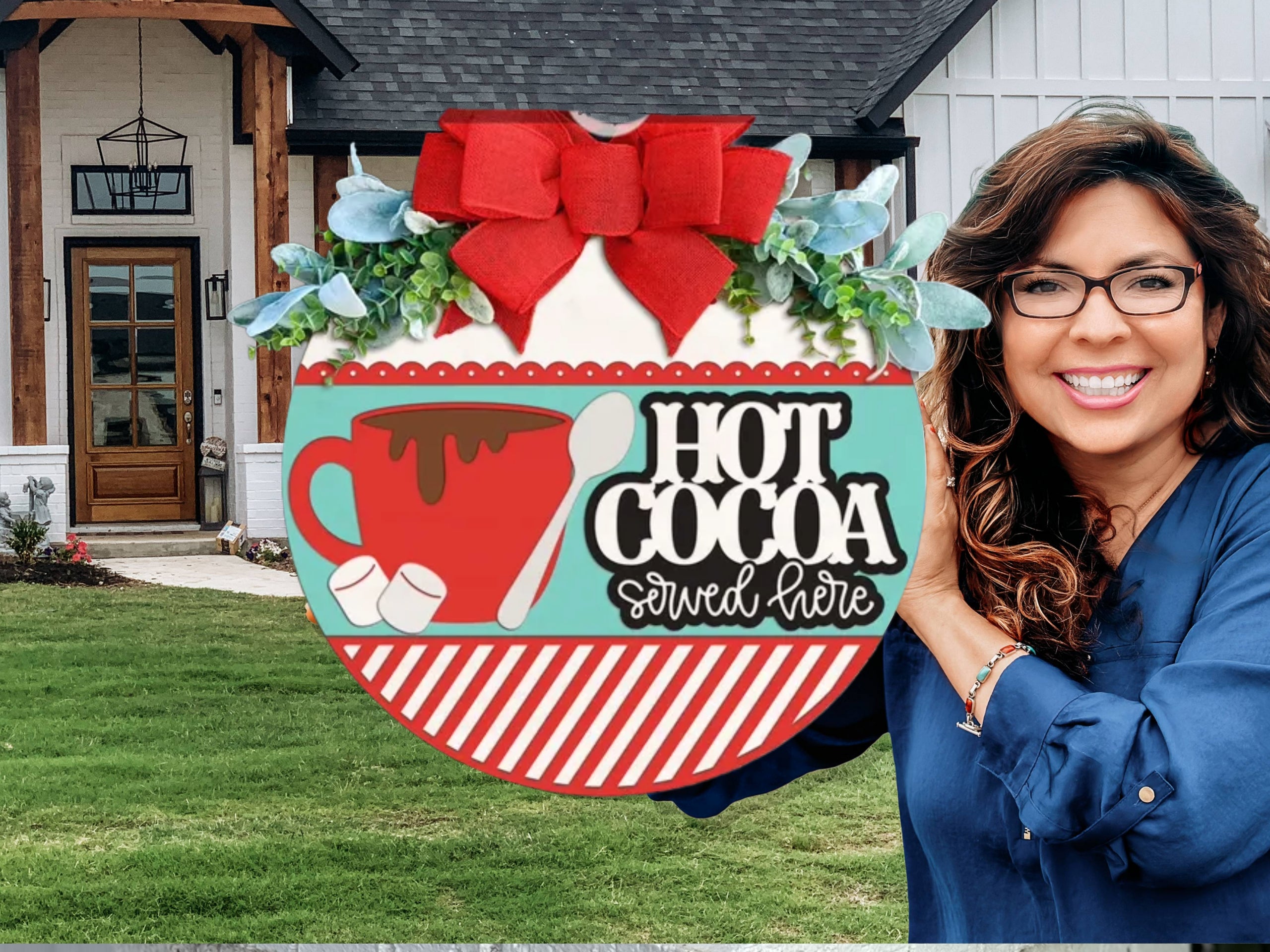 A decorative sign with a red cup, marshmallows, and a spoon reads “Hot Cocoa served here.” It features a large red bow, greenery, and a red-and-white striped border, sitting on a white brick ledge.