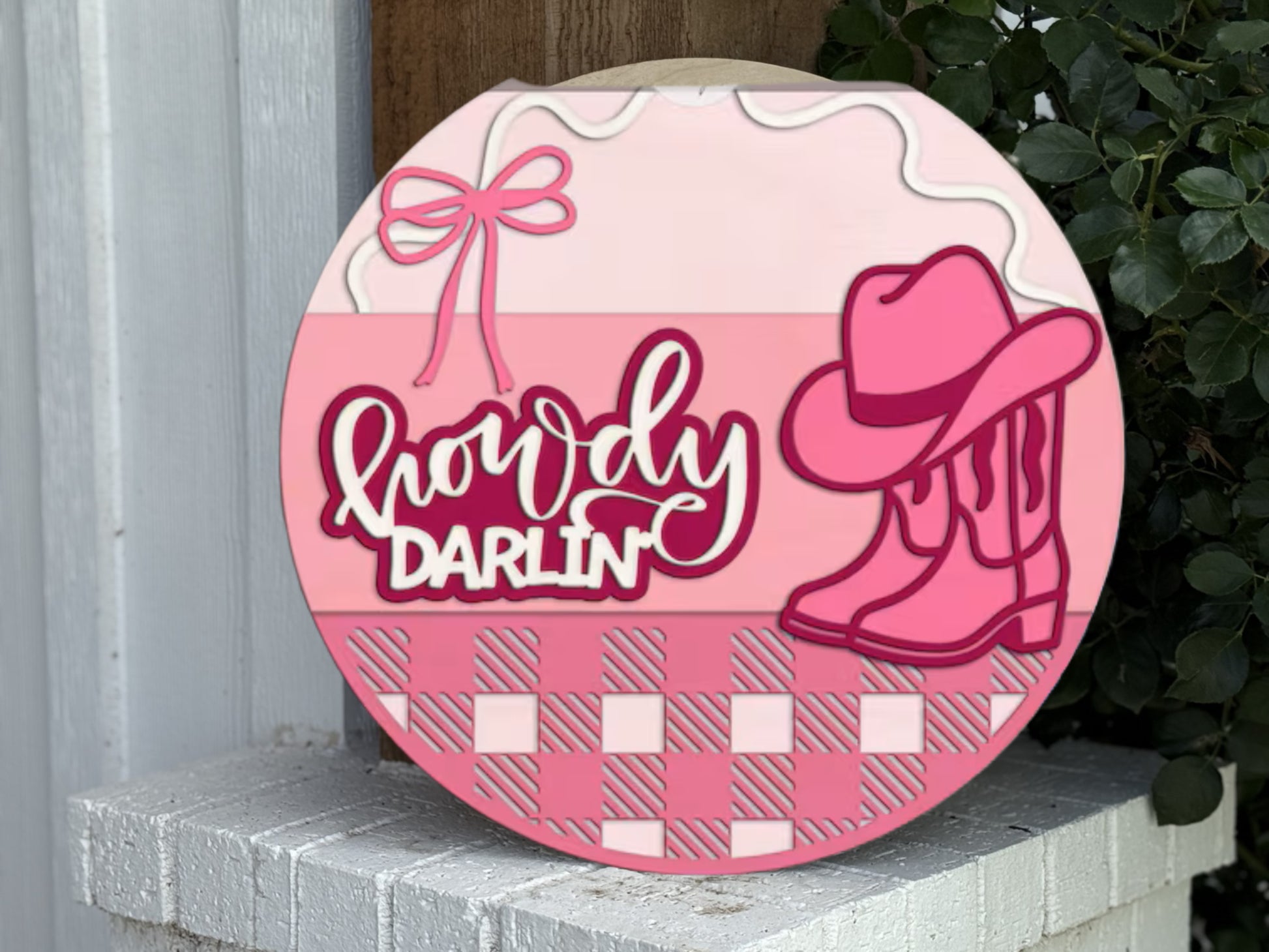 A round sign with Howdy Darlin’ in white and pink letters, decorated with a pink cowboy hat, boots, a bow, and a checkered pink pattern at the bottom, leaning against a white surface with green leaves nearby.