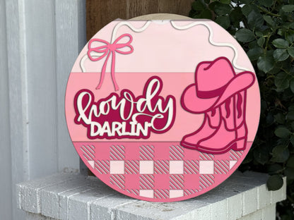 A round sign with Howdy Darlin’ in white and pink letters, decorated with a pink cowboy hat, boots, a bow, and a checkered pink pattern at the bottom, leaning against a white surface with green leaves nearby.