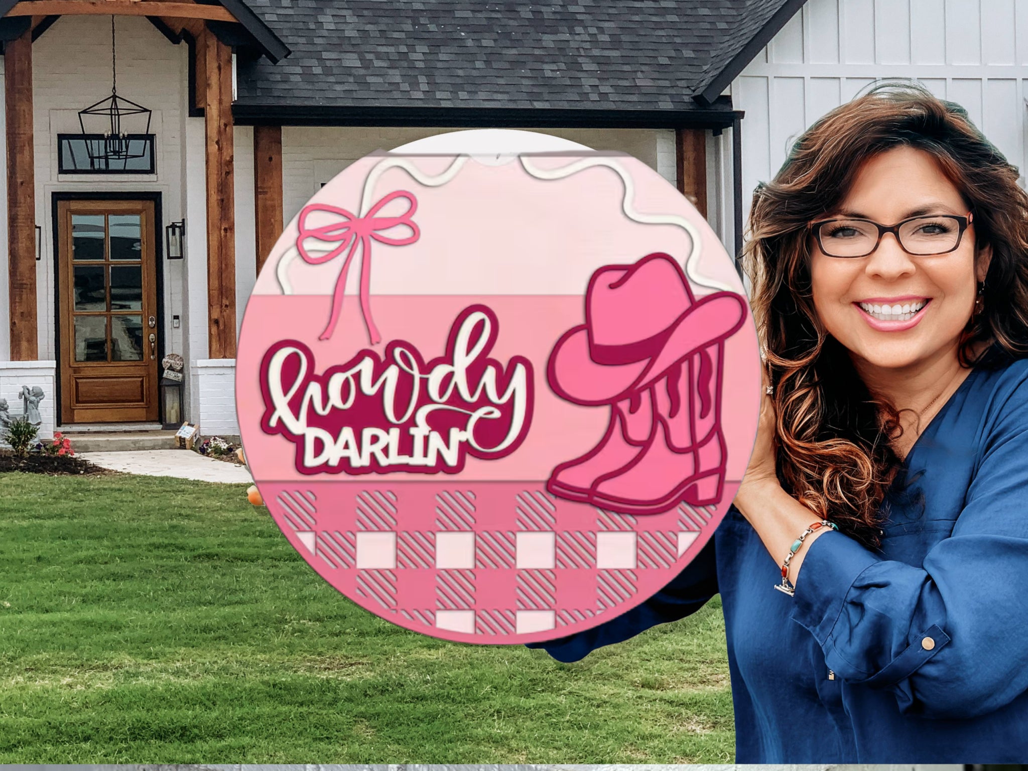 A smiling woman with long brown hair and glasses holds a round pink sign that says “Howdy Darlin’” with a cowboy hat and boots on it. She stands outside a modern house with wood and white siding.