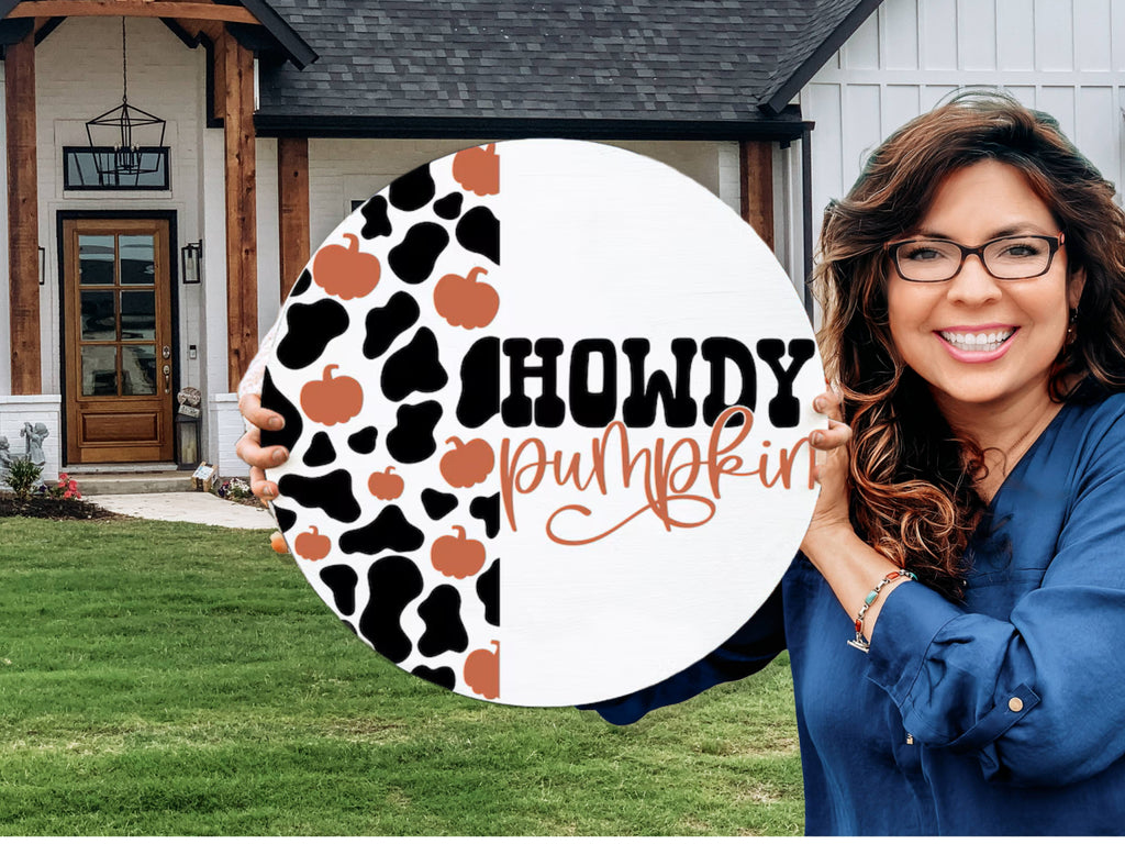 A smiling woman with long brown hair and glasses holds a round sign that says Howdy Pumpkin with a cow print and pumpkin pattern, standing in front of a modern house with a lawn.