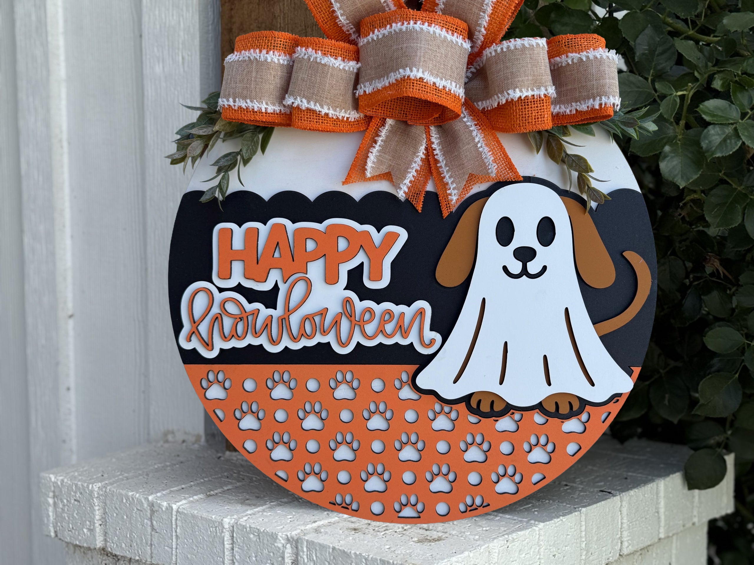 A round Halloween sign with a dog dressed as a ghost, orange and white paw prints, and “Happy Halloween” text, topped with an orange and burlap ribbon bow, displayed outside near green plants.