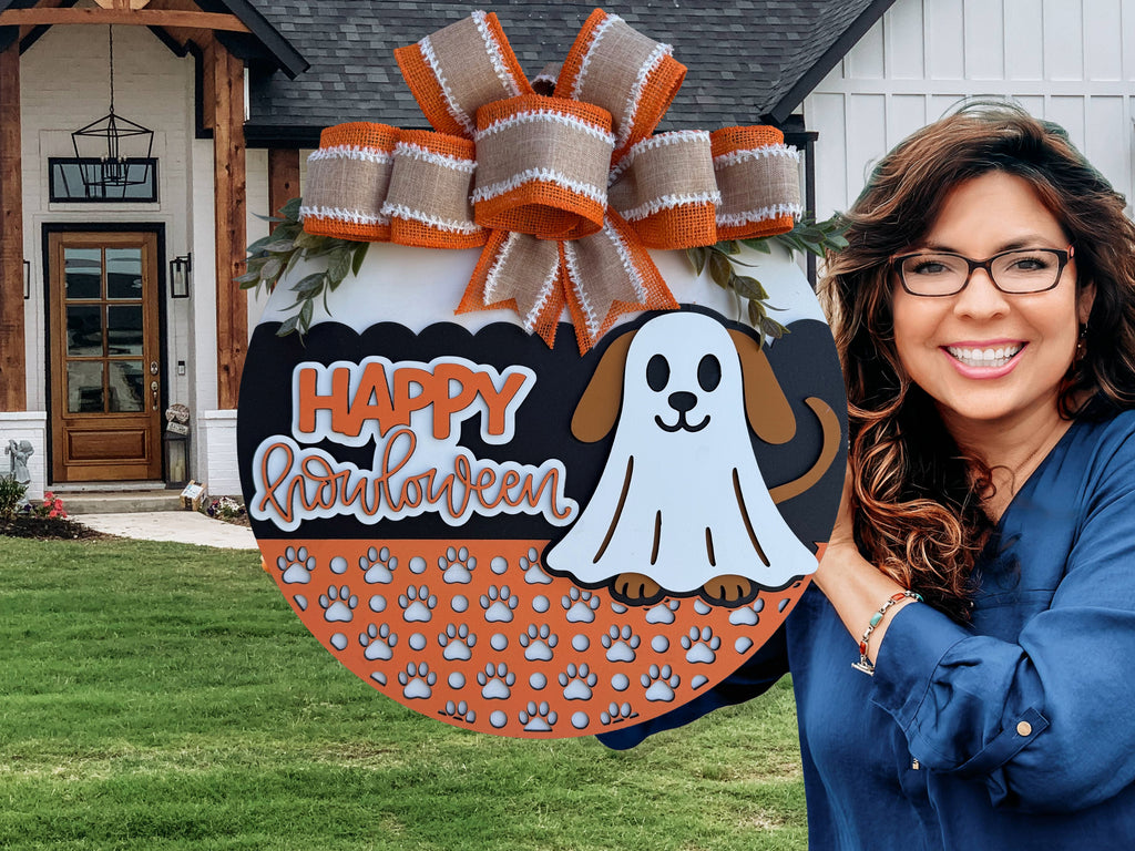 A smiling woman in glasses holds a Halloween sign with a cartoon dog in a ghost costume, orange and white bow, and “Happy Halloween” text. The background shows a modern house with a lawn.