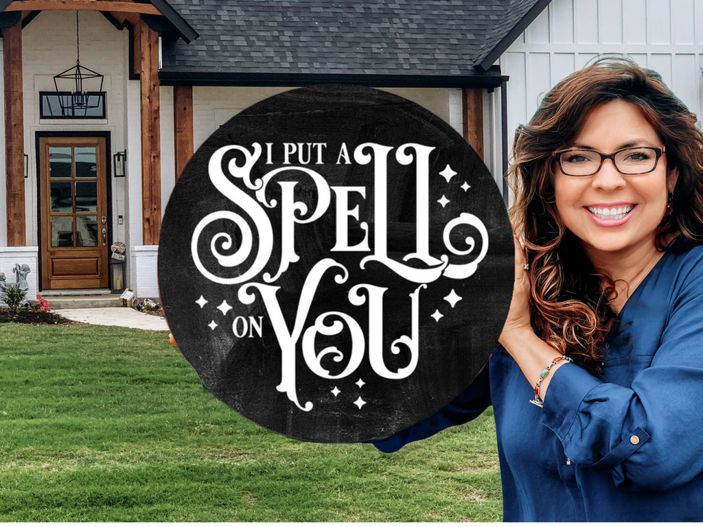 A smiling woman with long brown hair and glasses stands in front of a house, holding a round black sign that reads I PUT A SPELL ON YOU in decorative white lettering.