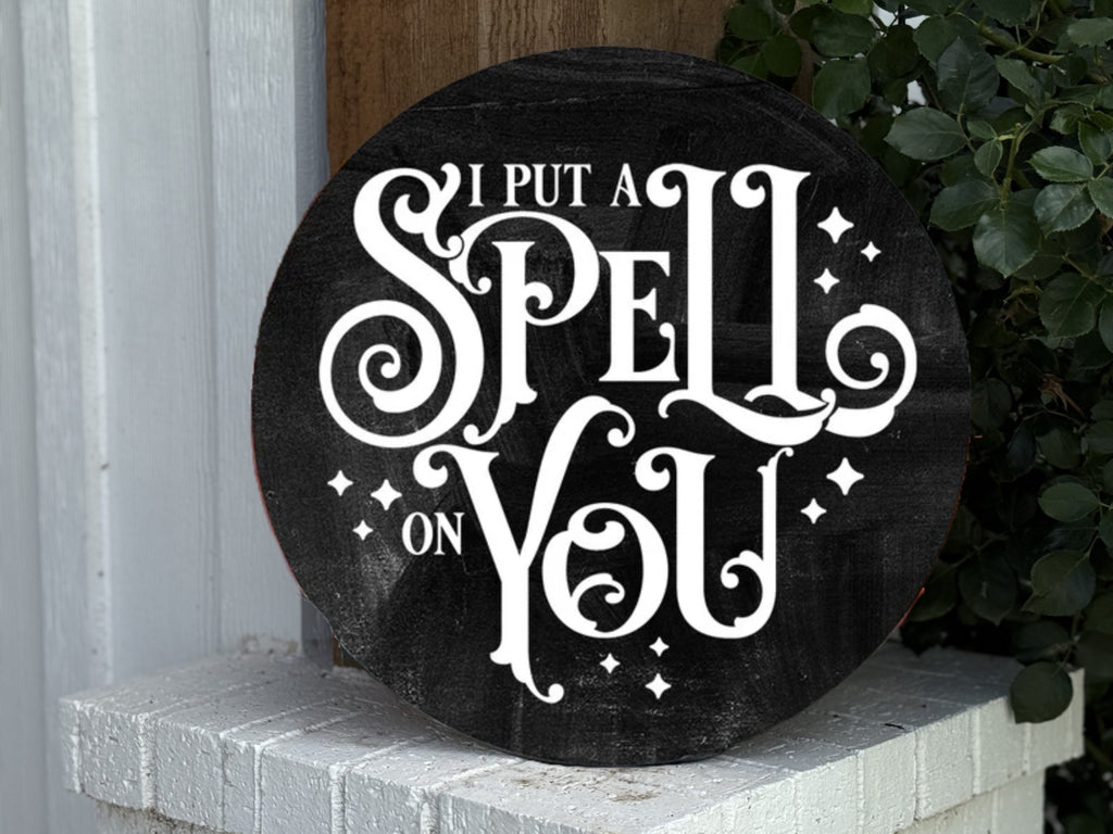 A round black sign with ornate white text reads, “I put a spell on you,” surrounded by small sparkling stars. The sign rests on a white brick surface beside green leaves and a wooden post.