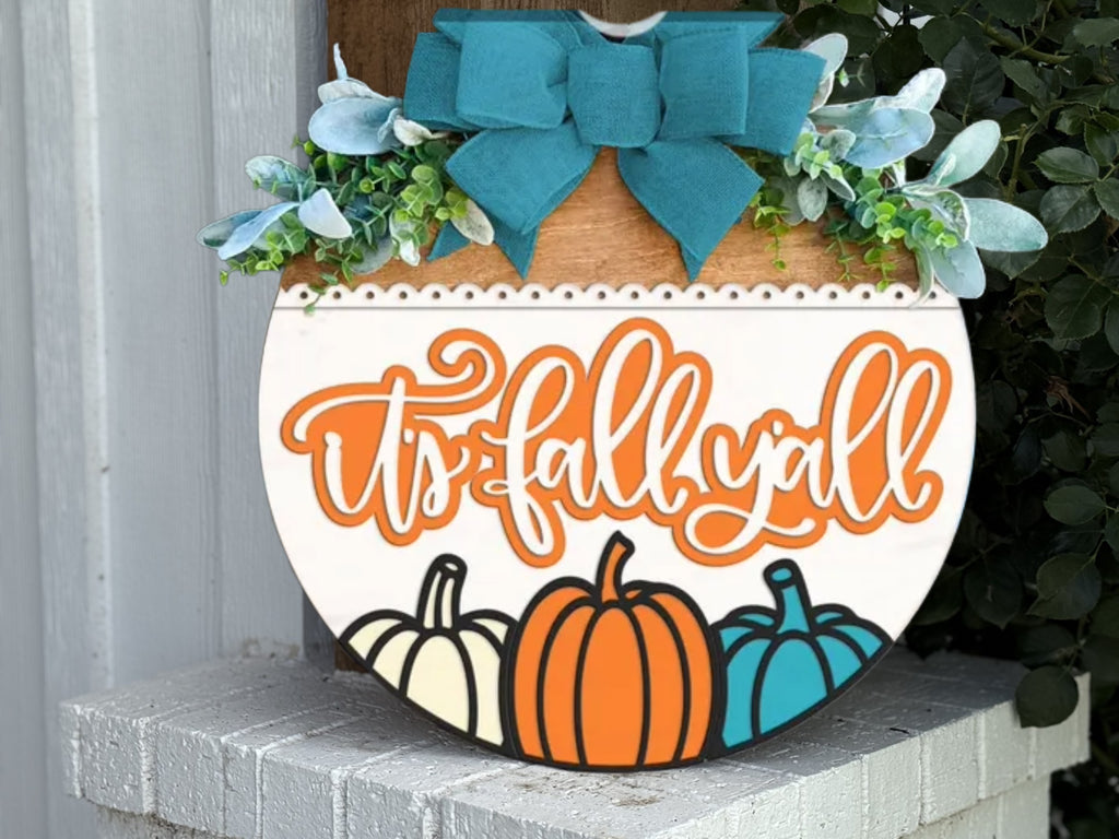 A woman with long brown hair and glasses smiles while holding a round sign that says hello FALL with a decorative pumpkin and autumn leaves, standing in front of a modern house.