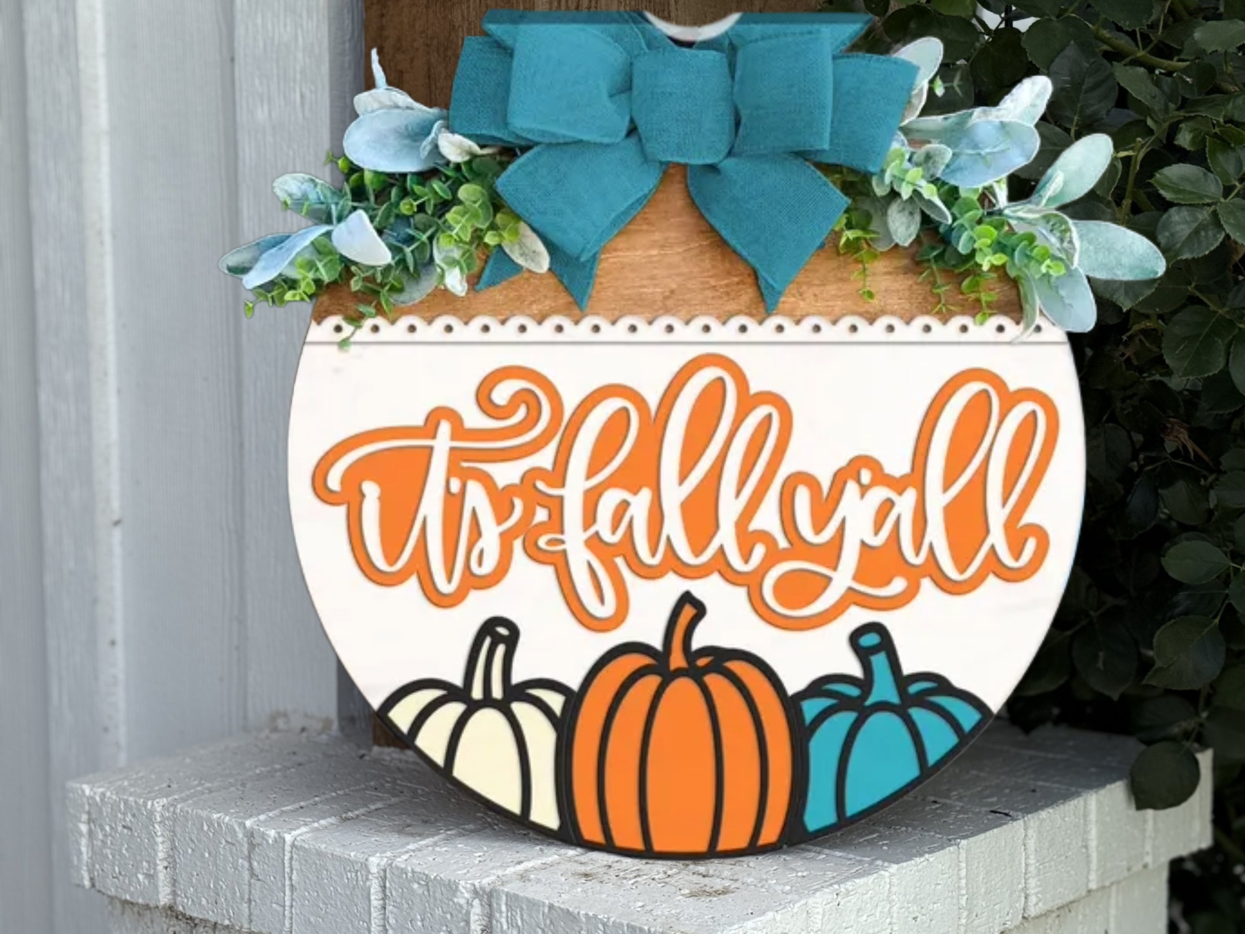 A woman with long brown hair and glasses smiles while holding a round sign that says hello FALL with a decorative pumpkin and autumn leaves, standing in front of a modern house.