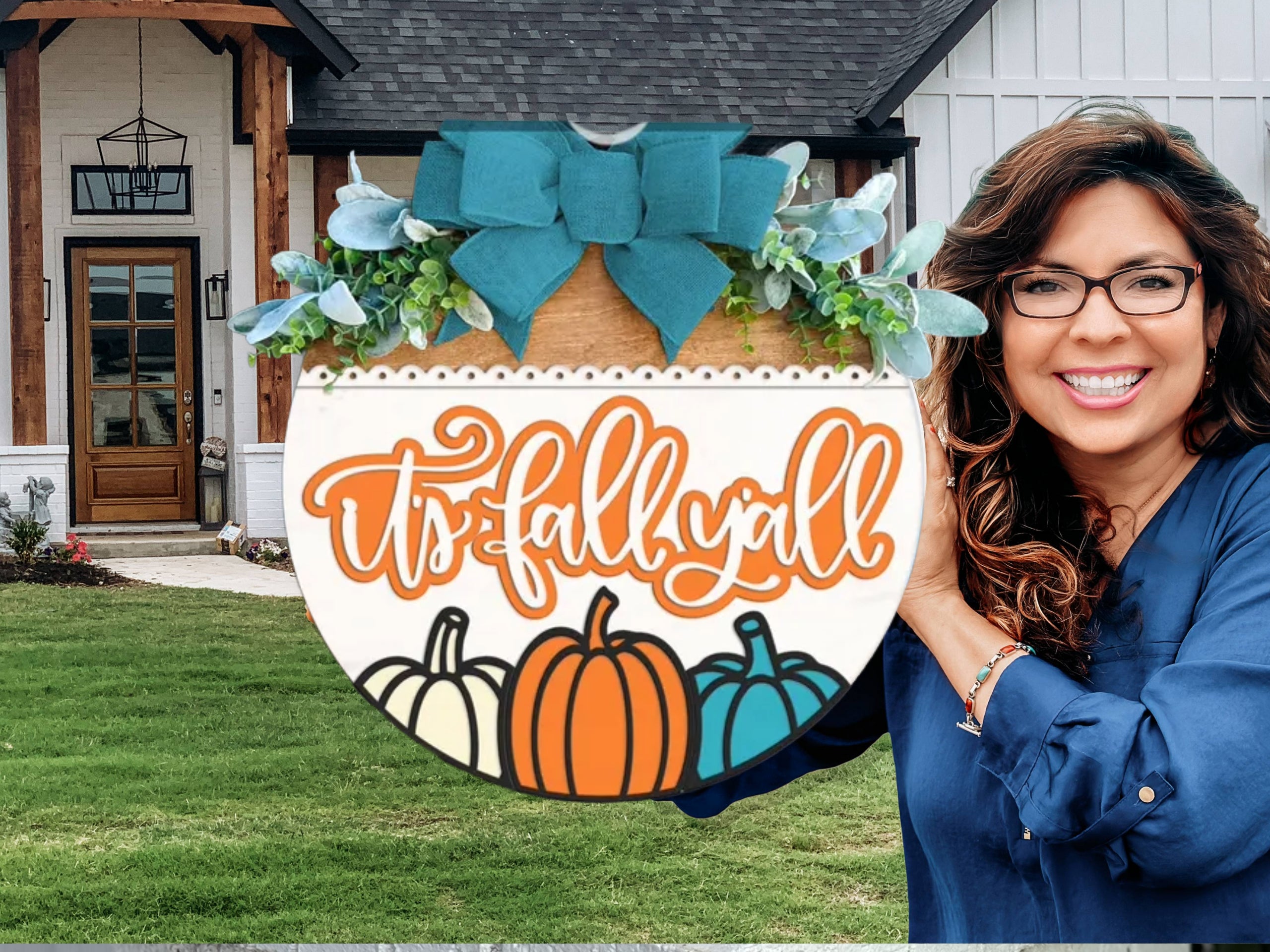 A woman with long brown hair and glasses smiles while holding a round sign that says hello FALL with a decorative pumpkin and autumn leaves, standing in front of a modern house.