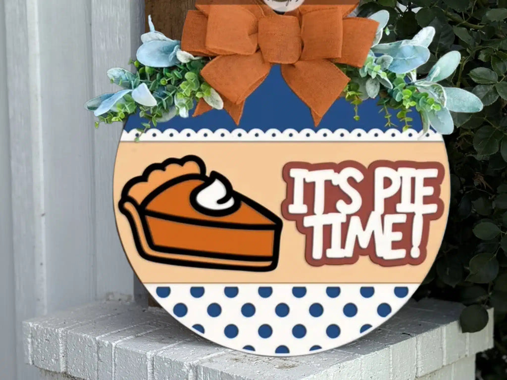 A round sign decorated with greenery and an orange bow reads ITS PIE TIME! next to a slice of pie with whipped cream. The bottom of the sign has a blue polka dot pattern.