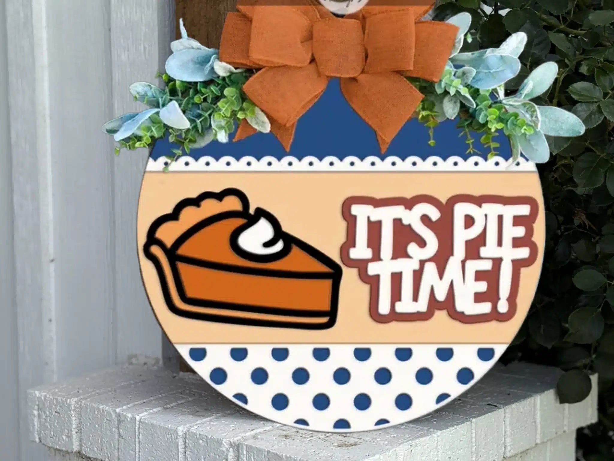 A round sign decorated with greenery and an orange bow reads ITS PIE TIME! next to a slice of pie with whipped cream. The bottom of the sign has a blue polka dot pattern.