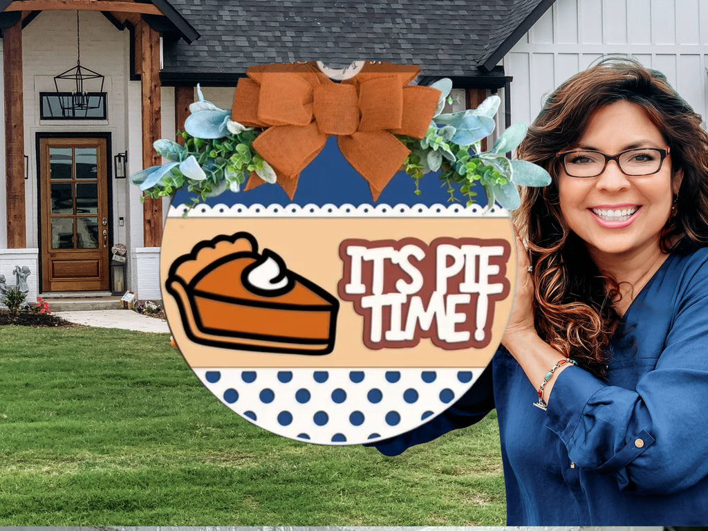 A smiling woman in glasses holds a round sign with a slice of pie, whipped cream, and the words ITS PIE TIME! in front of a house with brown and white exterior and green lawn.