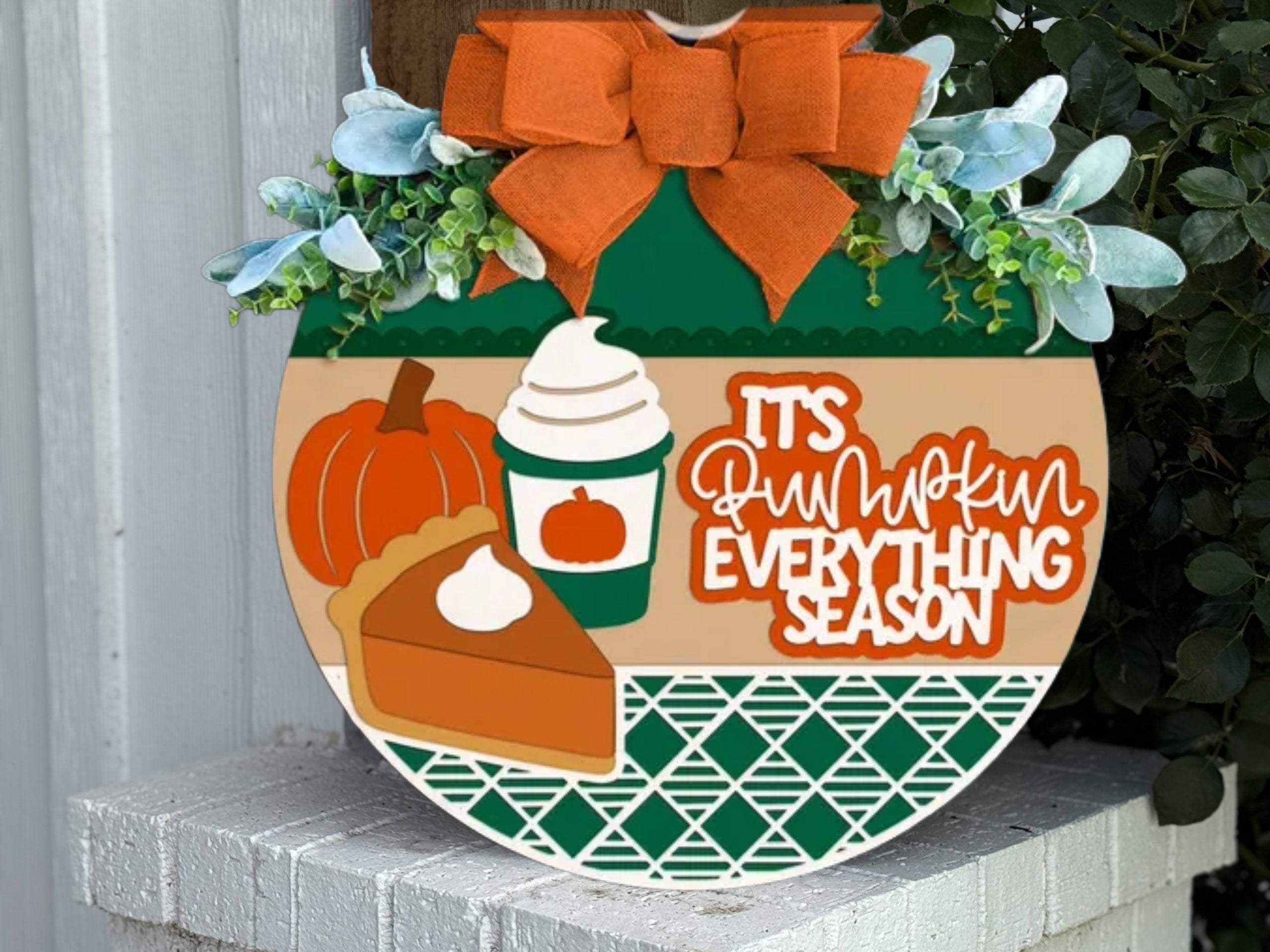 A decorative round sign with a pumpkin, pie slice, whipped drink, greenery, and an orange bow. The sign reads, Its pumpkin everything season. The background has green, beige, and geometric patterns.