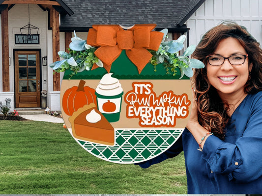 A decorative round sign with a pumpkin, pie slice, whipped drink, greenery, and an orange bow. The sign reads, Its pumpkin everything season. The background has green, beige, and geometric patterns.