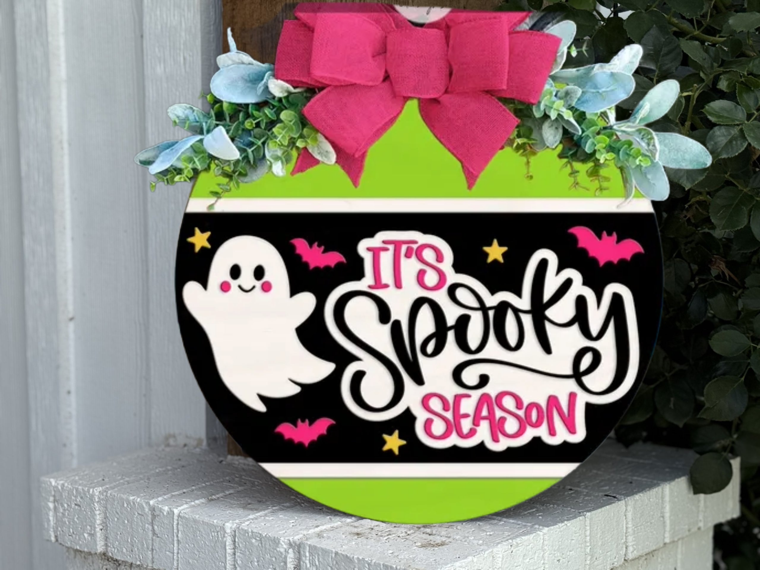 A round sign decorated with a big pink bow, greenery, and a design featuring a smiling ghost, bats, stars, and the words Its Spooky Season in bold letters. The sign is displayed outdoors on a white brick ledge.