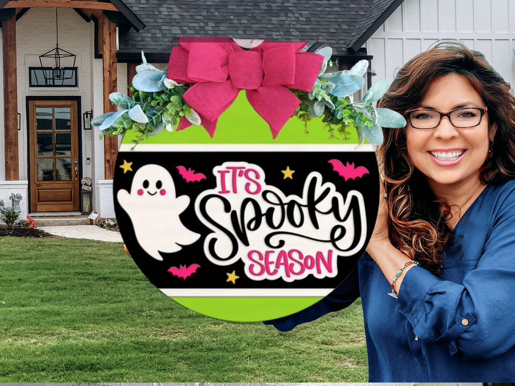 A round sign decorated with a big pink bow, greenery, and a design featuring a smiling ghost, bats, stars, and the words Its Spooky Season in bold letters. The sign is displayed outdoors on a white brick ledge.