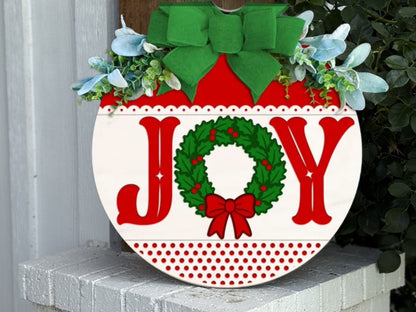 Circular holiday sign with the word JOY, where the O is a green wreath with a red bow. The sign is decorated with a large green bow and greenery at the top, and rests on a white brick ledge.