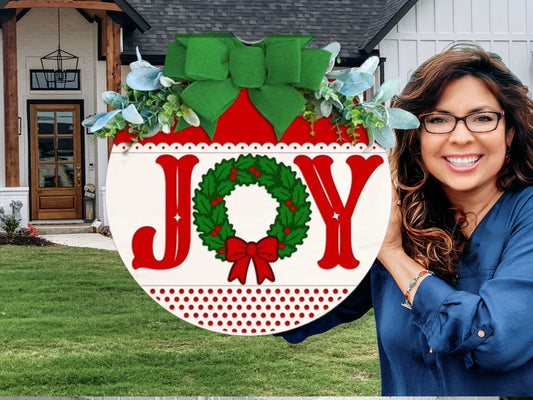 Circular holiday sign with the word JOY, where the O is a green wreath with a red bow. The sign is decorated with a large green bow and greenery at the top, and rests on a white brick ledge.