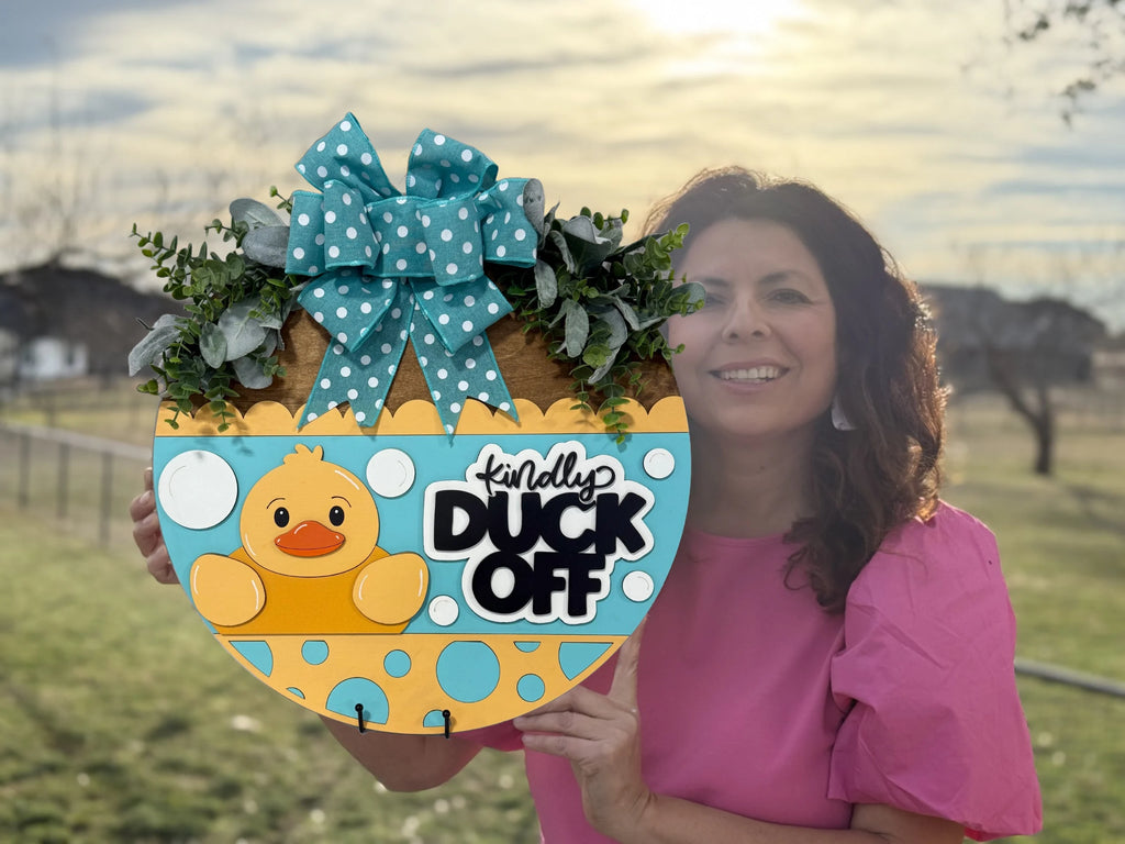A woman in a pink shirt holds a decorative, duck-themed sign outdoors. The sign features a yellow duck and reads Kindly Duck Off, adorned with a blue polka dot bow and greenery. The background shows a grassy area and a partly cloudy sky.