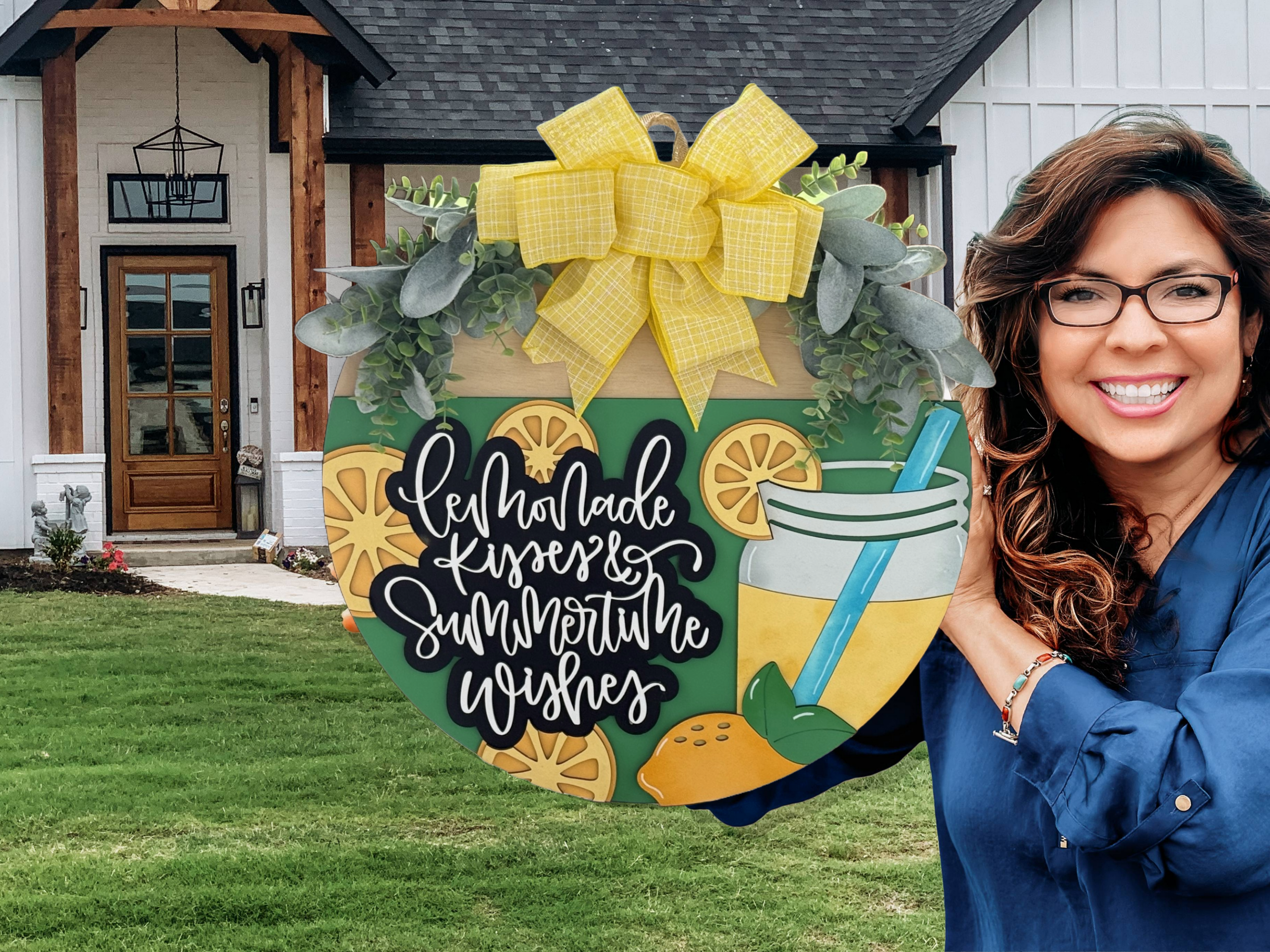 A smiling woman with long brown hair and glasses holds a large decorative sign reading “Lemonade Kisses & Summertime Wishes,” featuring lemons, a lemonade drink, greenery, and a yellow plaid bow, standing in front of a modern farmhouse.