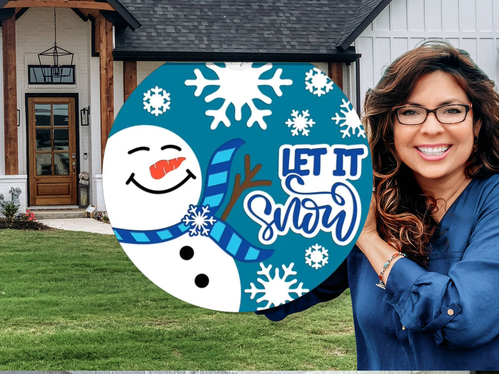 A smiling woman holds a round sign with a snowman, snowflakes, and the words Let It Snow in front of a modern house with a lawn.