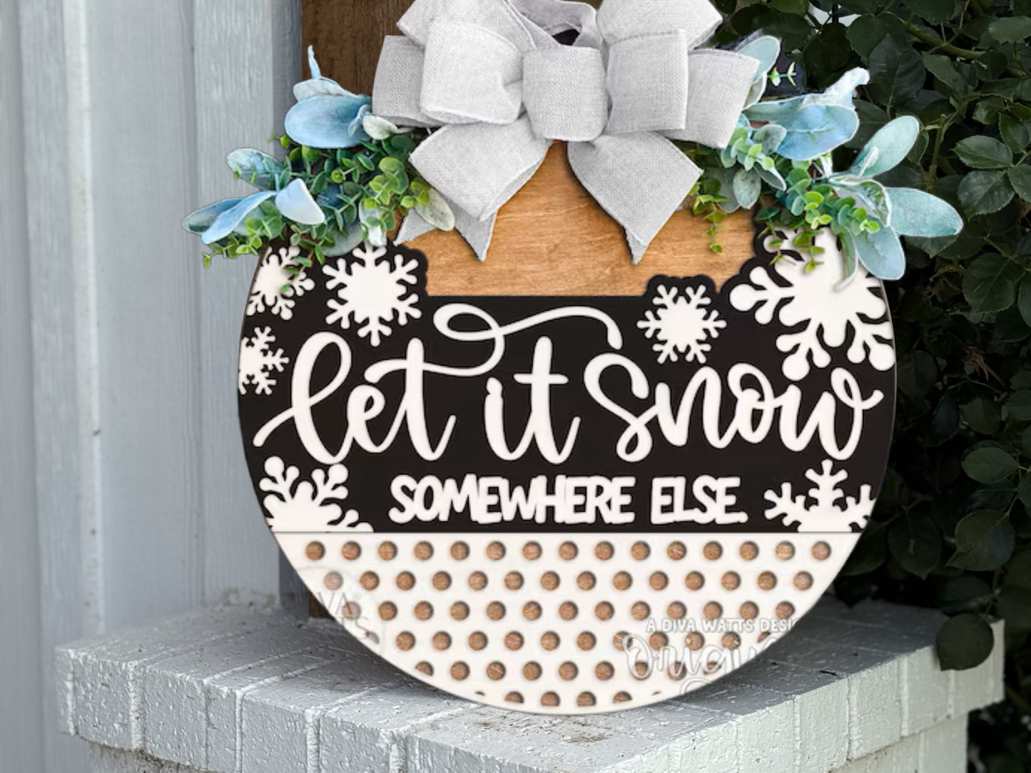 A round decorative sign with a bow, greenery, snowflakes, and text that reads Let it snow, somewhere else, leaning against a white painted brick surface.