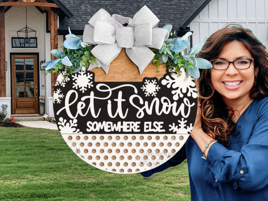A smiling woman holds a decorative round sign with a bow, greenery, and the text Let it snow somewhere else in front of a modern farmhouse entrance.