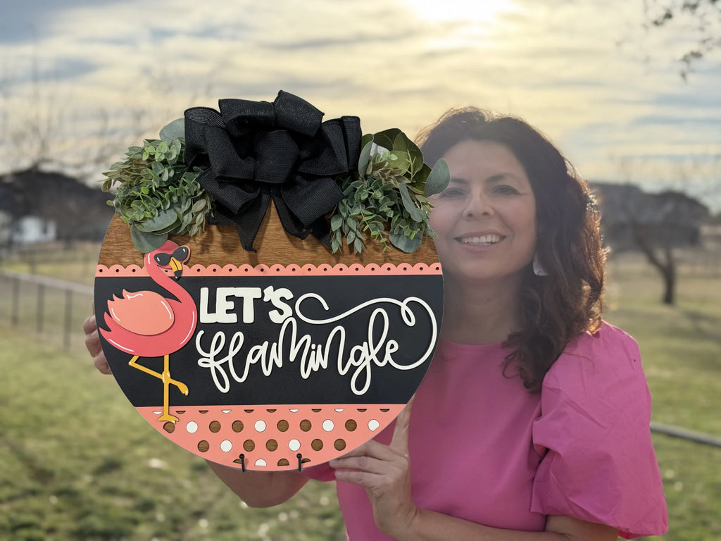 A woman in a pink blouse holds a decorative round sign outdoors. The sign features a pink flamingo illustration, the text Lets Flamingle, and is adorned with a black bow and greenery. The background shows a grassy area and a cloudy sky.