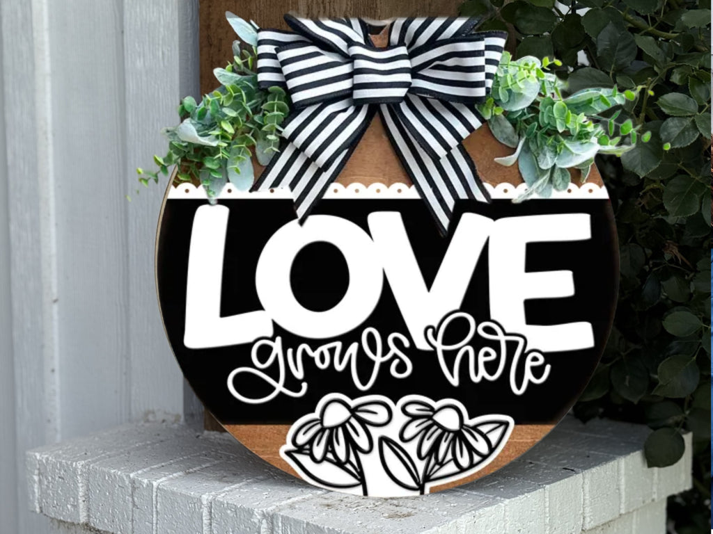 A round wooden sign with the words LOVE grows here, decorated with a black and white striped bow, greenery, and white floral designs, leaning against a white brick wall.
