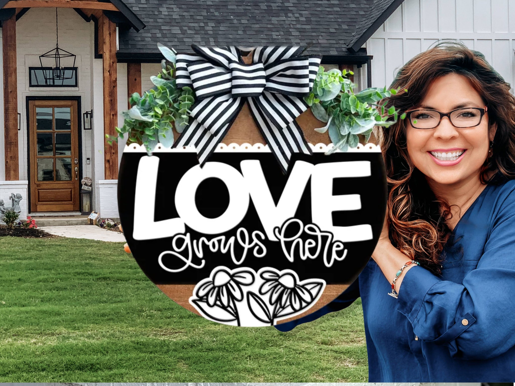 A smiling woman with long brown hair and glasses stands in front of a modern house, holding a round sign with a black and white bow that reads LOVE grows here with floral decorations.