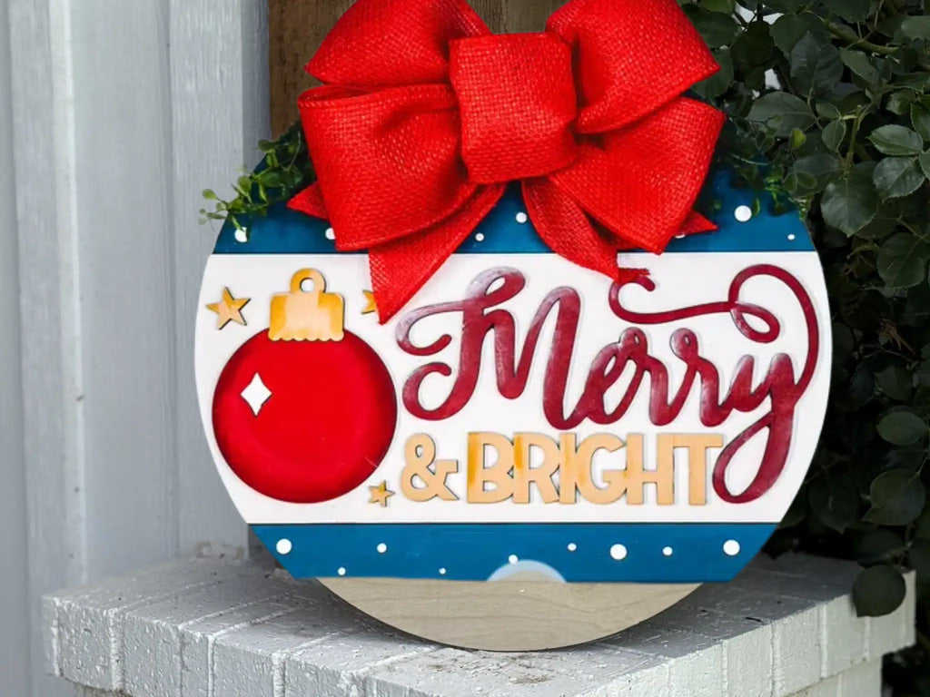 A festive round sign with a large red bow, a red Christmas ornament, and the words Merry & Bright in decorative letters, displayed on a white brick surface with green leaves nearby.