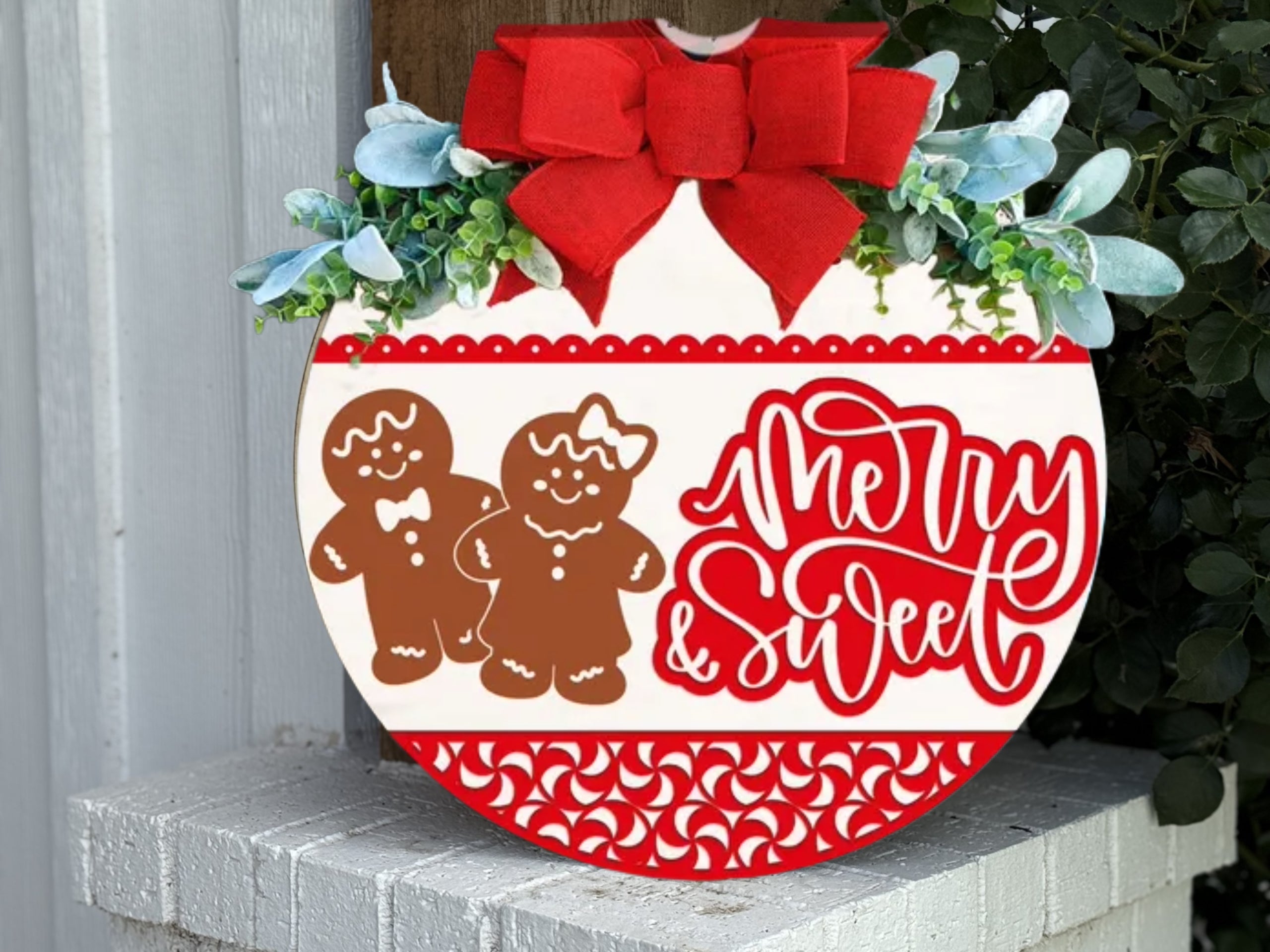 A round Christmas sign decorated with a red bow, greenery, and two gingerbread figures. The sign reads Merry & Sweet in red script and has red decorative patterns along the top and bottom edges.