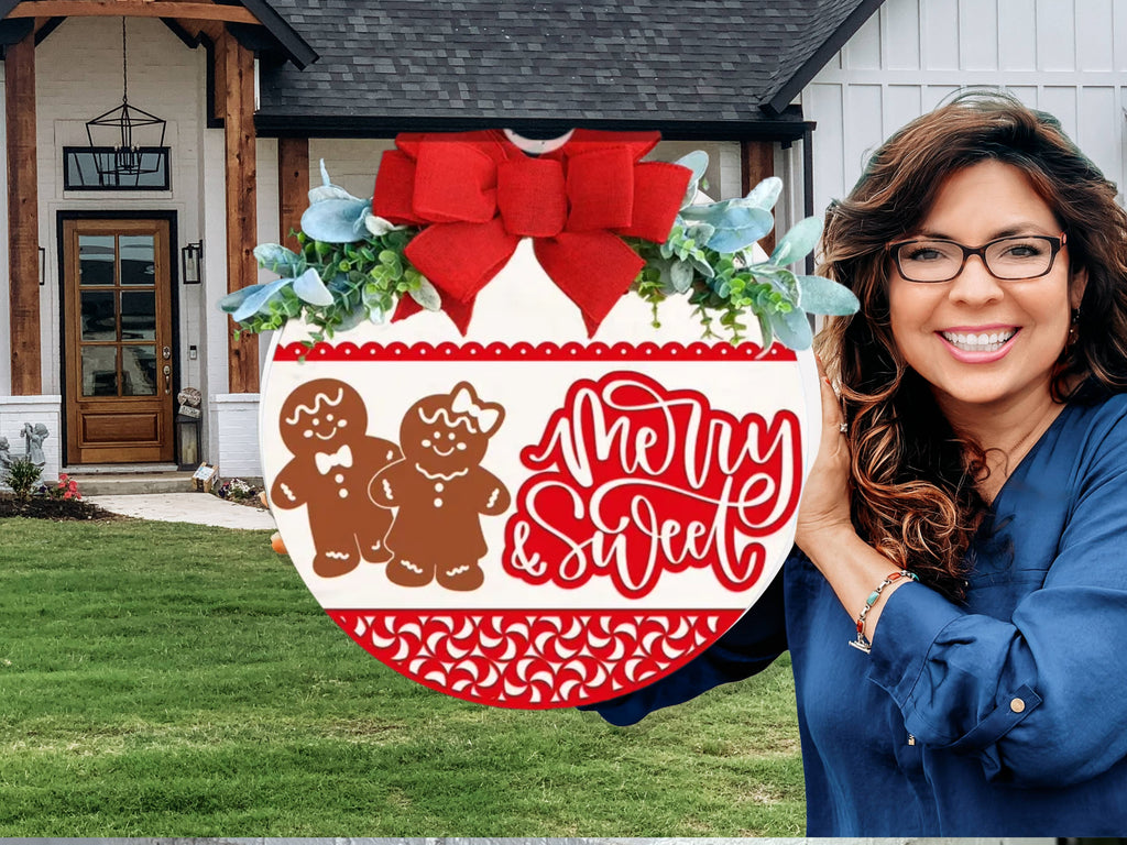 A smiling woman with long brown hair and glasses holds a festive sign featuring gingerbread figures, greenery, a red bow, and the words “Merry & Sweet” in front of a modern house with a green lawn.