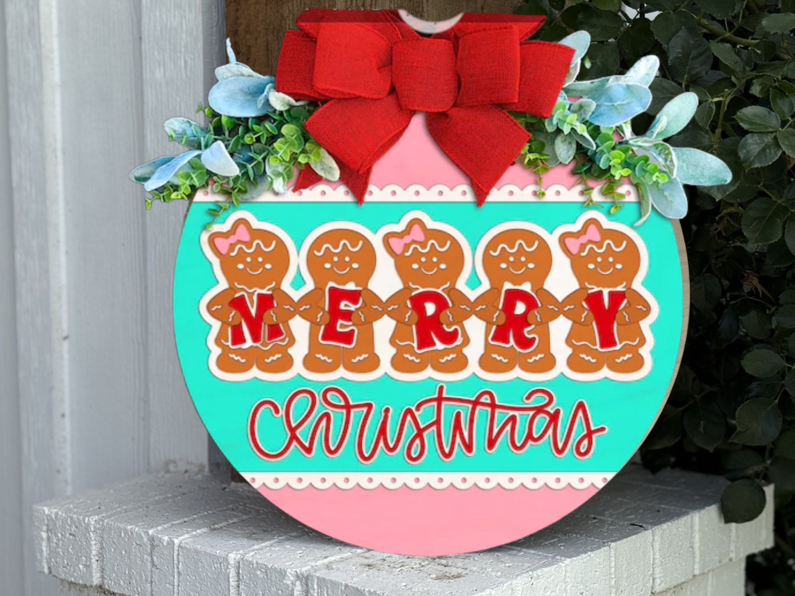 A round, festive sign with a red bow, greenery, and gingerbread people spelling MERRY above the word Christmas in cursive, hangs on a white brick wall corner.