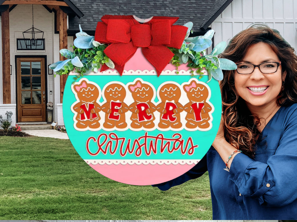 A smiling woman in glasses holds a round Christmas sign decorated with gingerbread people, greenery, and a large red bow. The sign says MERRY CHRISTMAS. She stands outside a modern house with a green lawn.