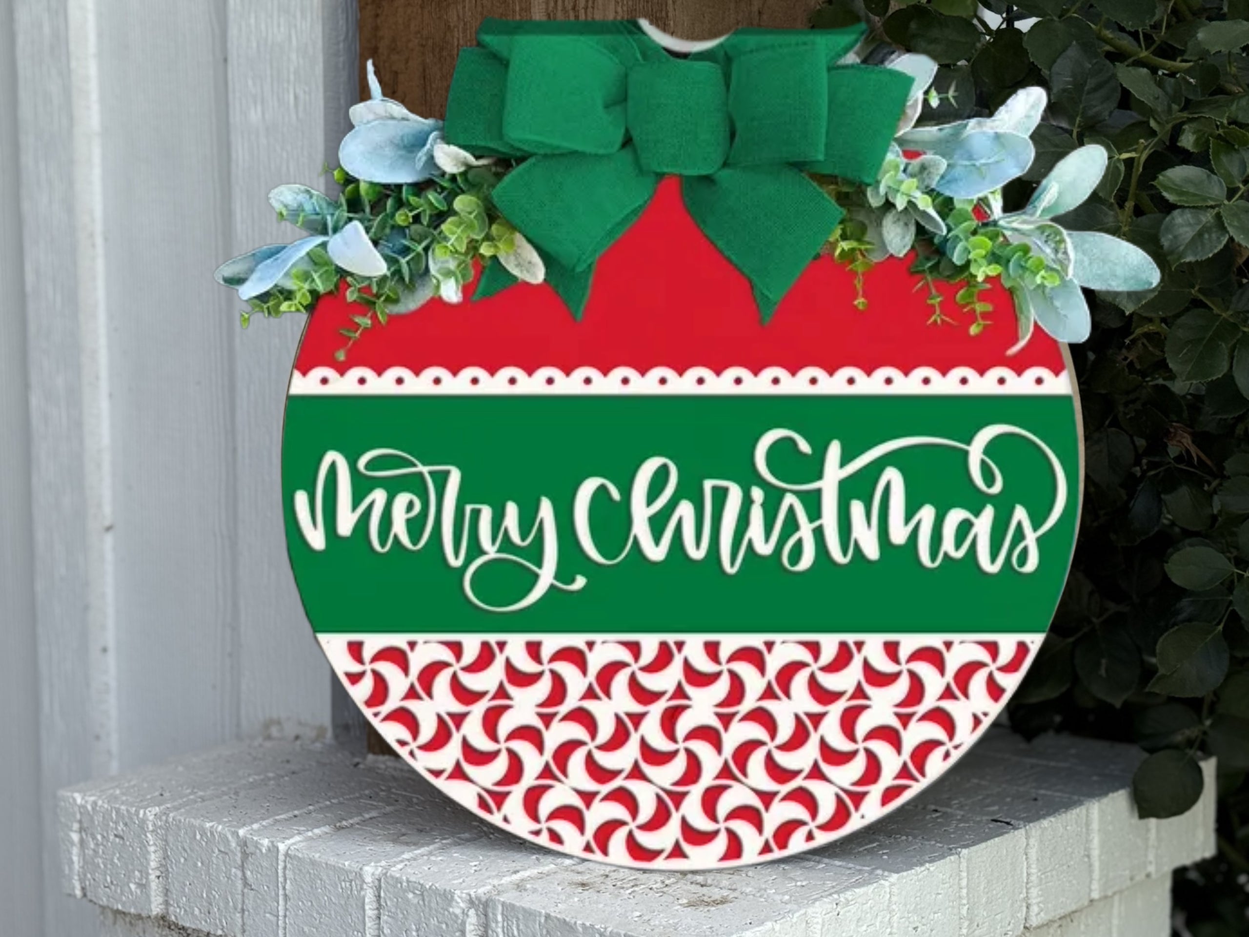 A round Christmas sign with a green bow, white and blue flowers, and greenery on top. The sign says Merry Christmas in white script on green, with red and white peppermint patterns below.