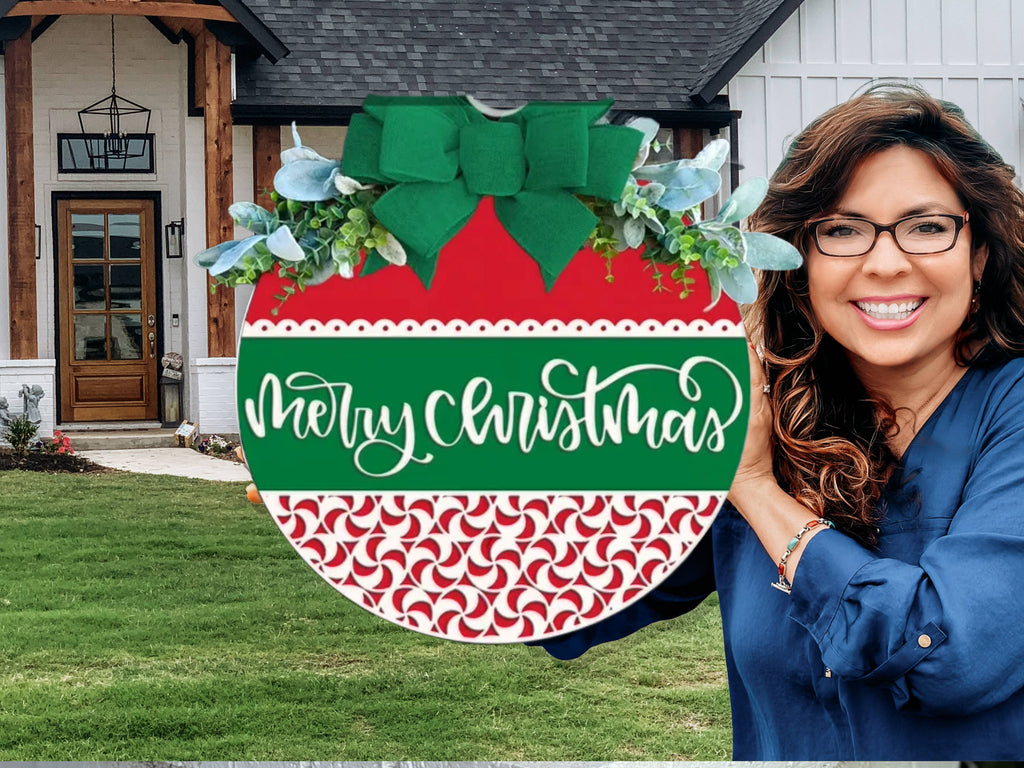 A smiling woman with long brown hair and glasses holds a large festive ornament sign that reads Merry Christmas in front of a modern house with white and wood accents.